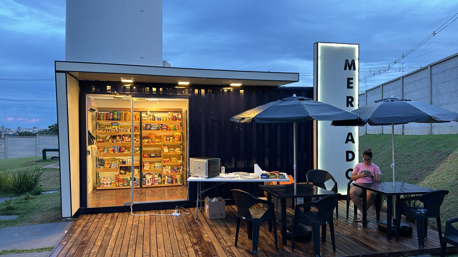 Loja Omnimart em container iluminada ao anoitecer com deck e mesas